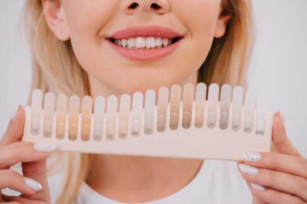 cropped view of smiling woman holding teeth color palette isolated on white, teeth whitening concept